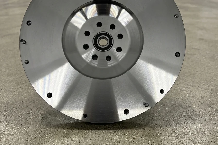 Close-up macro photograph of a 5-axis CNC milling machine cutting the central bore of a heavy custom sand-cast ductile iron flywheel. Sharp metal shavings are flying off the cutting tool, and bright white liquid coolant is spraying onto the dark gray iron surface. The background is a blurred industrial factory setting.